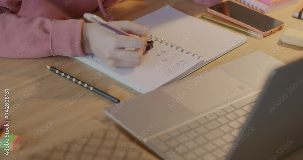 Close-up of hand writing numbers in notebook while schoolgirl doing mathematics homework at desk at home. Education and exam preparation concept.