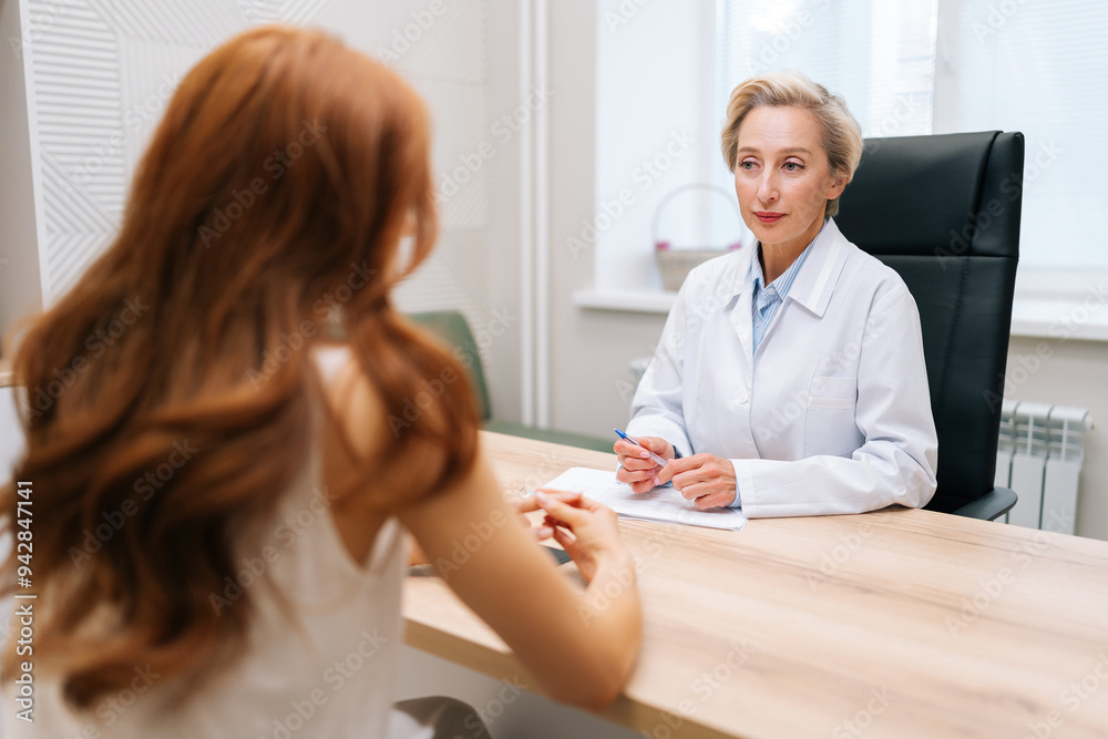 Focused mature adult female portioner therapist giving professional advice, health recommendation to young unrecognizable redhead patient. Concept of treatment, chronic disease prevention, healthcare.