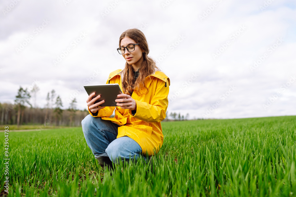 A young farmer woman in a green field, intently using a tablet. She wears a yellow jacket and glasses, enjoying the serene, cloudy day outdoors. The concept of technology, gardening. A young farmer woman in a green field, intently using a tablet. She wears a yellow jacket and glasses, enjoying the serene, cloudy day outdoors. The concept of technology, gardening.