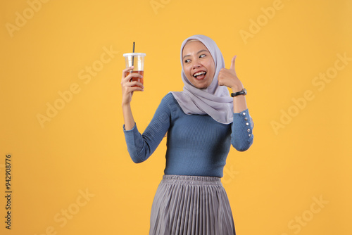 happy asian indonesian muslim woman holding and drinking ice tea in plastic cup and giving thumb up finger gesture on isolated yellow background photo