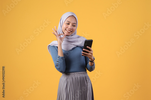 happy asian indonesian muslim woman holding smart phone giving ok finger gesture on isolated yellow background