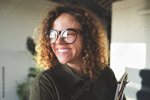 Young businesswoman with a big smile standing in an office. She has a clipboard in her arms, curly hair and wears big black glasses.