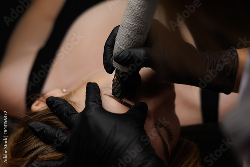 A dark cosmetology office performs precise permanent eyebrow makeup work using a tattoo tool. Permanent eyebrow makeup, eyebrow tattooing with brown pigment for PMU.