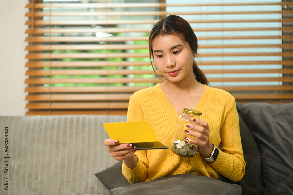 Young woman holding glass jar filled with coins and attentively looking at a bank passbook. Budgeting or saving money concept Young woman holding glass jar filled with coins and attentively looking at a bank passbook. Budgeting or saving money concept