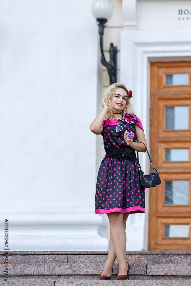 Blonde girl in a bright dress and sunglasses on the street near a building on a walk on a summer day. Hipster Blonde girl in a bright dress and sunglasses on the street near a building on a walk on a summer day. Hipster