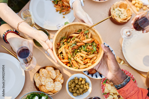 Hands passing a bowl of pasta at a table with various dishes, seen from above. Diverse group enjoying a meal of mixed cuisine, featuring pasta, olives, and crackers