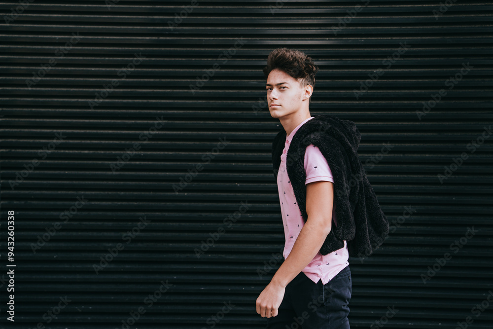 young man wearing a pink polo shirt, walking with a jacket over his shoulder against a black, ribbed wall. Concept of casual fashion and urban style young man wearing a pink polo shirt, walking with a jacket over his shoulder against a black, ribbed wall. Concept of casual fashion and urban style
