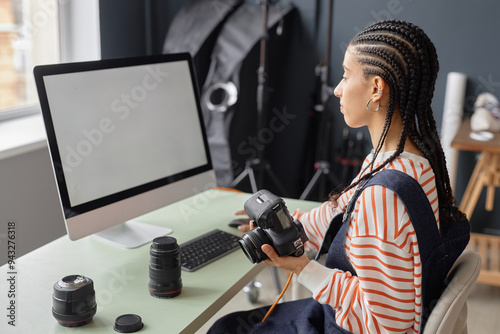 Side view portrait of Black young woman with braided hair as female photographer using computer with blank white screen in photo studio and holding camera photo