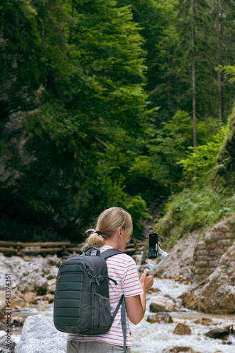 woman with backpack shoots video or takes photos on smartphone in wooded area. She stands near a mountain stream, surrounded by dense greenery and stones. The landscape creates an atmosphere of tranqu