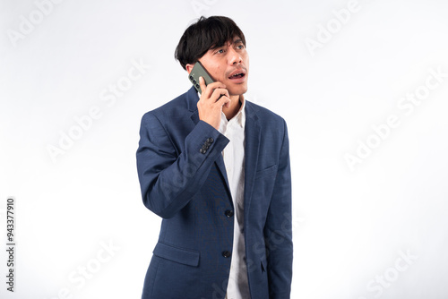 A surprised Asian businessman in a navy blue suit holds a smartphone to his ear, reacting with shock during a phone call. The white background emphasizes his expression