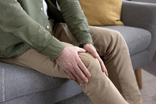 Middle-aged man holding knee in discomfort while sitting on sofa. Concept of joint pain, aging, health issues. Image captures emotion of concern regarding personal wellness and mobility challenges.