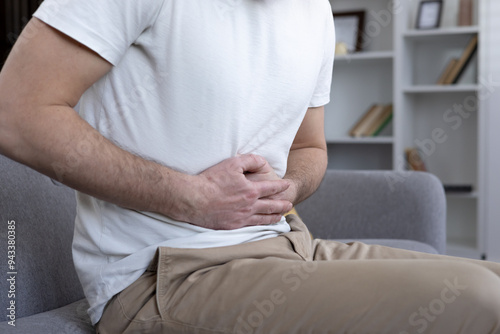 Man holding stomach in discomfort, indicating abdominal pain. Clad in casual attire, seated on sofa in modern living room. Captures concept of health issues, illness, or indigestion.