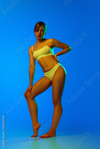Full-length image of attractive, beautiful young girl with skinny, slim body posing in underwear against blue studio background in neon. Concept of female beauty, body and skin care, sport, health