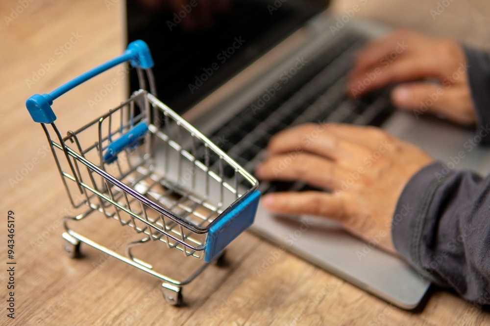 Close-up of mini shopping trolley and hands typing on laptop. Online shopping concept.