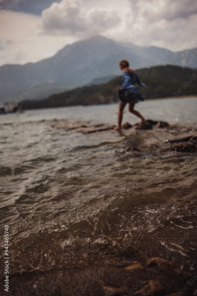 Fototapeta premium A person walks barefoot on rocky shoreline during golden hour against a backdrop of mountains