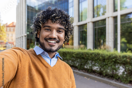 Man with curly hair taking selfie in urban area. Wearing orange sweater over blue shirt. Smiling broadly, enjoying outdoor setting. Technology emphasizes friendly and approachable nature.