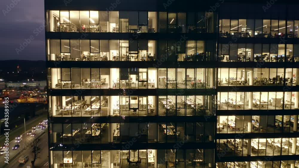 A panoramic drone shot captures illuminated windows a modern glass skyscraper, where business professionals are seen working late into the night, reflecting the non-stop nature urban corporate life. A panoramic drone shot captures illuminated windows a modern glass skyscraper, where business professionals are seen working late into the night, reflecting the non-stop nature urban corporate life.