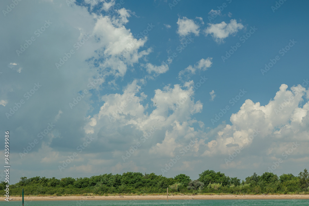 vista dettagliata di una piccola porzione di costa, con una spiaggia e un bosco verde, sotto un cielo azzurro, parzialmente nuvoloso, di giorno, in estate