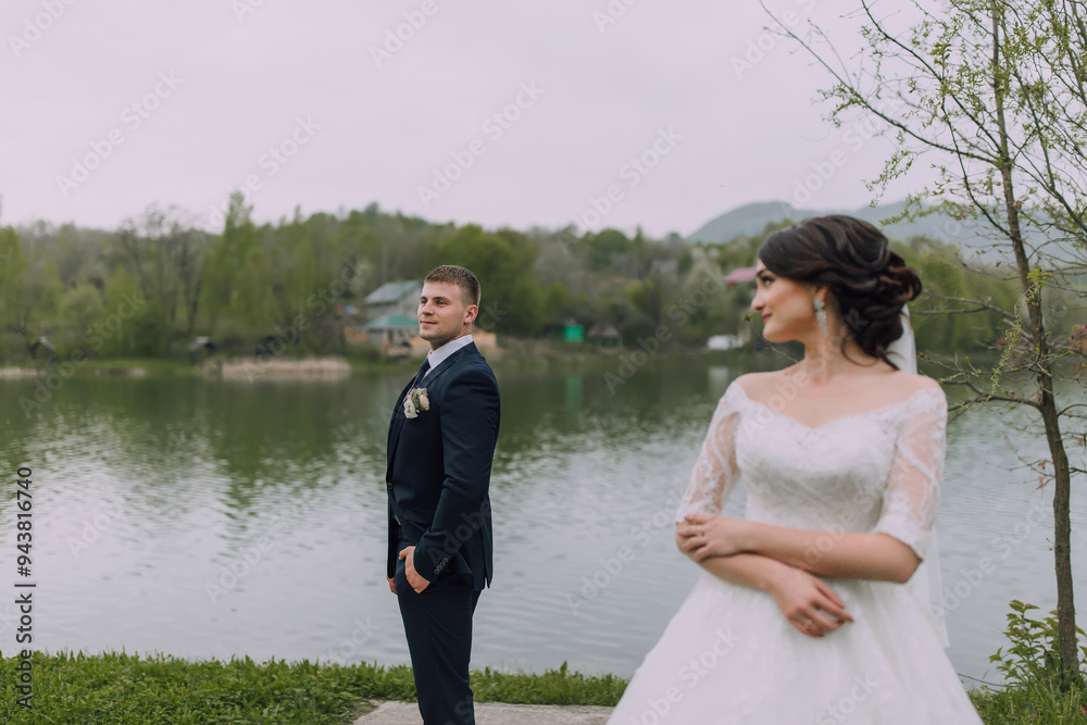 A bride and groom stand on a grassy bank overlooking a lake. The bride is wearing a white dress and a veil, while the groom is dressed in a suit. The scene is serene and romantic A bride and groom stand on a grassy bank overlooking a lake. The bride is wearing a white dress and a veil, while the groom is dressed in a suit. The scene is serene and romantic
