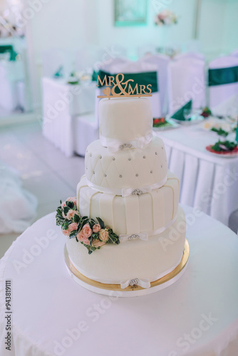 A four tier wedding cake with a white base and a pink and white floral design. The cake is topped with a gold and white ribbon and the words "Mr. And Mrs." are written on the top of the cake