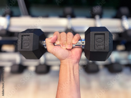 hand gripping dumbbell with a strong, determined hold. The background features a blurred view of other gym equipment, highlighting the focus on the dumbbell and the intensity of the workout, fitness