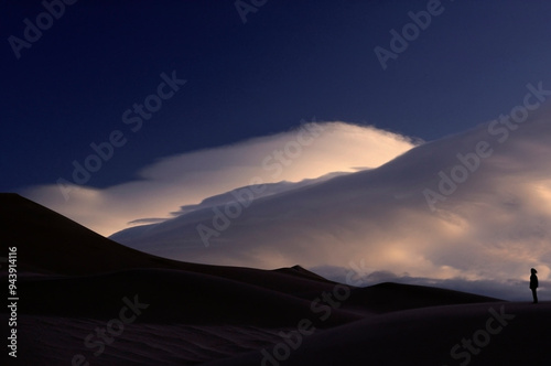 Silhouetted figure stands on the vast sand dunes as cloud formations roll overhead in Great Sand Dunes National Park And Preserve in Colorado, USA; Colorado, United States of America