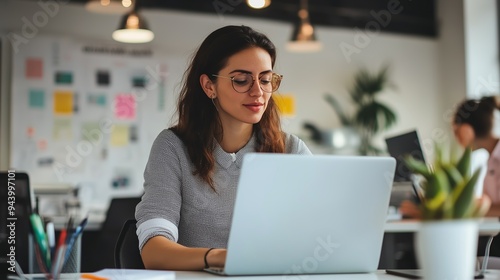 Young woman working on a laptop at a modern office during the day while colleagues engage in various tasks in the background