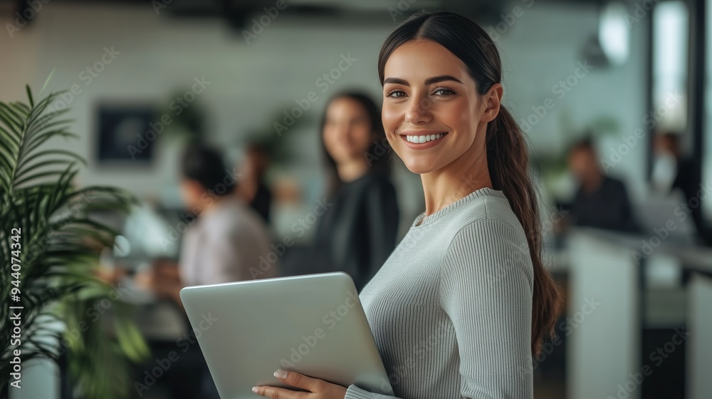 A young woman smiles confidently while holding a laptop in a vibrant coworking space during a busy workday A young woman smiles confidently while holding a laptop in a vibrant coworking space during a busy workday