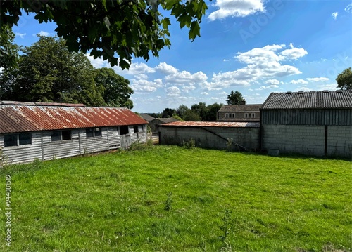 A grassy yard is surrounded by old, weathered buildings with corrugated roofs. Trees and blue sky with fluffy clouds are visible in the background near, Dewsbury, Yorkshire, UK photo