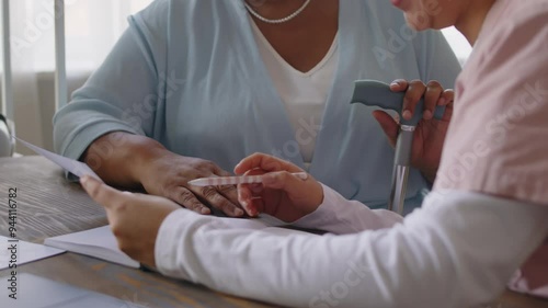 Female caregiver with pen and document discussing paper with unrecognisable senior woman holding cane and putting hand on chest while sitting at desk