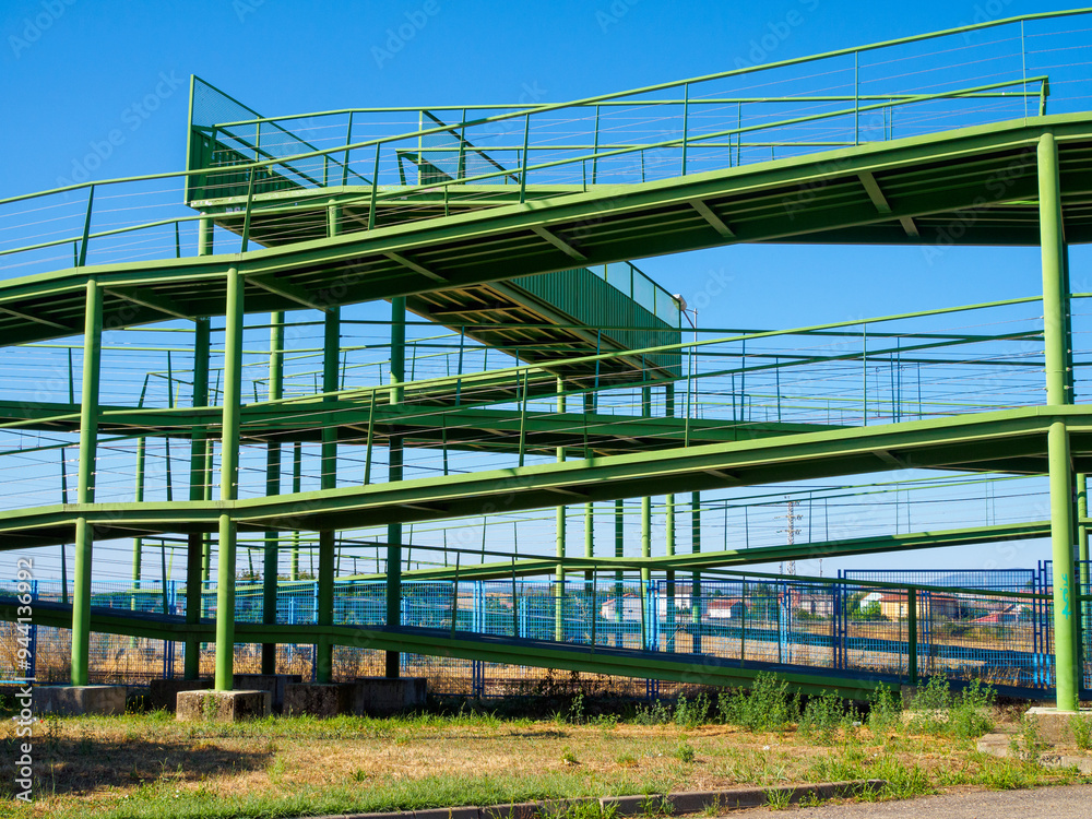 A complex green metal framework with geometric patterns and multiple levels serves as an industrial overpass, bridging a road and railway tracks, offering a unique view of the surrounding urban area A complex green metal framework with geometric patterns and multiple levels serves as an industrial overpass, bridging a road and railway tracks, offering a unique view of the surrounding urban area