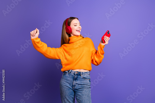 Photo of cheerful teenager girl in orange turtleneck and jeans carefree dancing with smartphone earphones isolated on purple color background