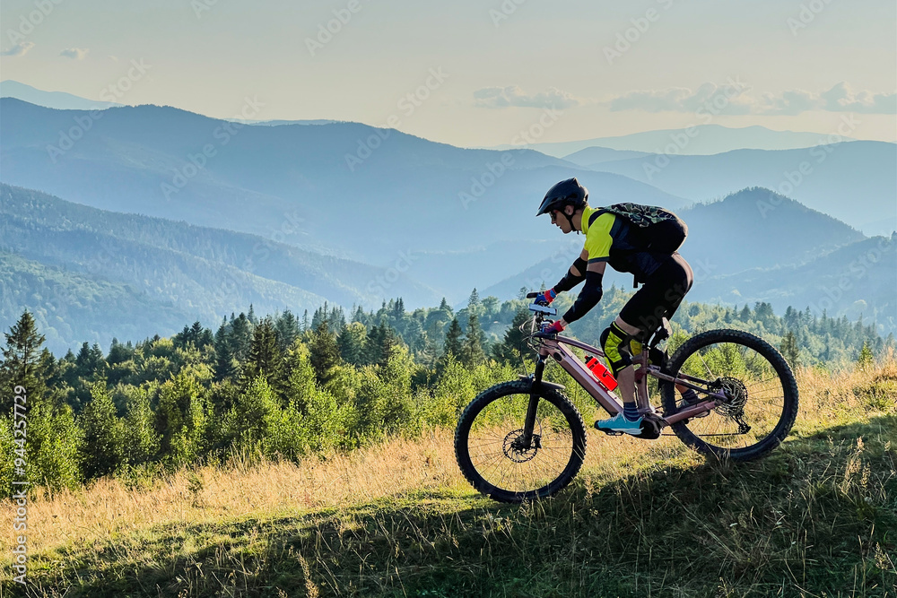 Cyclist man riding electric mountain bike outdoors. Male tourist biking along grassy trail in the mountains, wearing helmet and backpack. Concept of sport, active leisure and nature. Cyclist man riding electric mountain bike outdoors. Male tourist biking along grassy trail in the mountains, wearing helmet and backpack. Concept of sport, active leisure and nature.