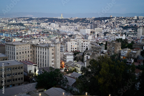 The densely packed city of Algiers begins to be lit at evening time, with the Monument to Martyrs on the horizon and mountains behind.