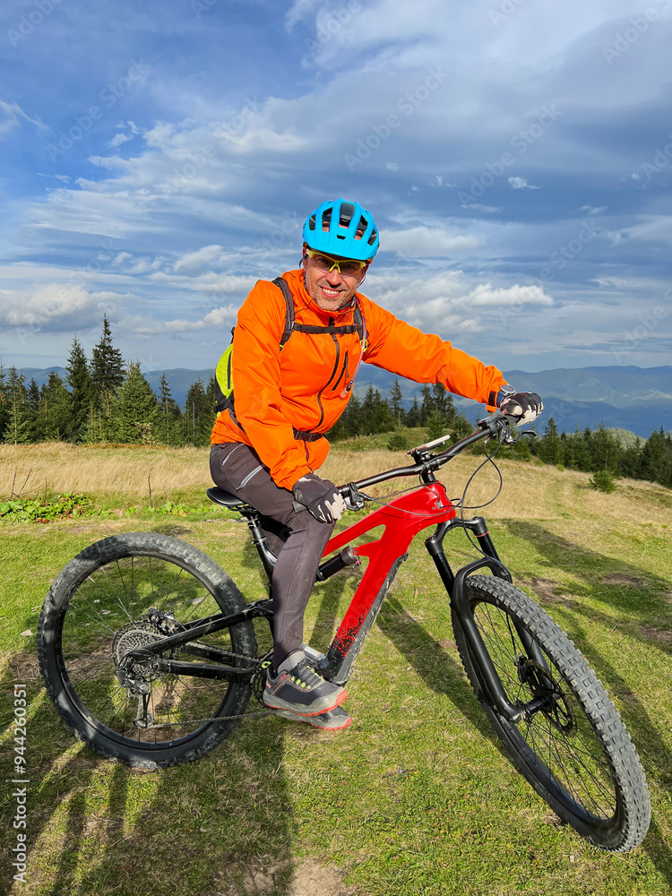 Cyclist man riding electric bike outdoors on sunny day. Portrait of smiling male tourist resting on grassy hill, enjoying beautiful mountain landscape, wearing helmet. Concept of active leisure. Cyclist man riding electric bike outdoors on sunny day. Portrait of smiling male tourist resting on grassy hill, enjoying beautiful mountain landscape, wearing helmet. Concept of active leisure.