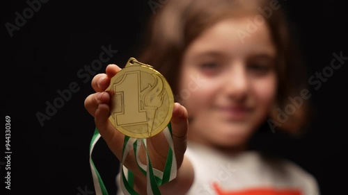 A schoolgirl proudly extends a first-place gold medal towards the camera, showcasing her achievement.