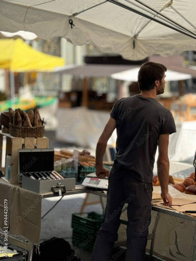 A vendor at an outdoor market standing behind a stall filled with freshly baked bread, preparing to serve customers under a canopy. The atmosphere is lively and focused on artisanal goods A vendor at an outdoor market standing behind a stall filled with freshly baked bread, preparing to serve customers under a canopy. The atmosphere is lively and focused on artisanal goods