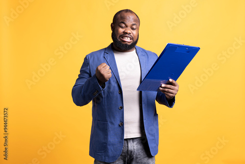 Man celebrating while browsing though financial report pages, happy with good job, studio background. Joyous worker exalting while reading paperwork, satisfied with good profits turnover