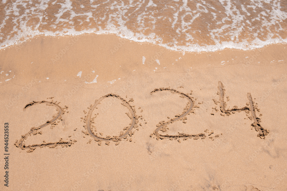 Fototapeta premium Looking ahead: The year '2024' emerges in the sand as the ocean waves softly approach, symbolizing the dawn of a new chapter.