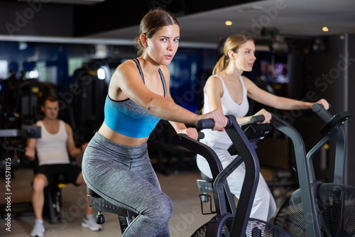 Girl spends time in gym, improves her physical shape with exercises. Female sport studio hall visitor uses exercise air bike to pump leg muscles. Strength and motivation, sport, fitness goal photo