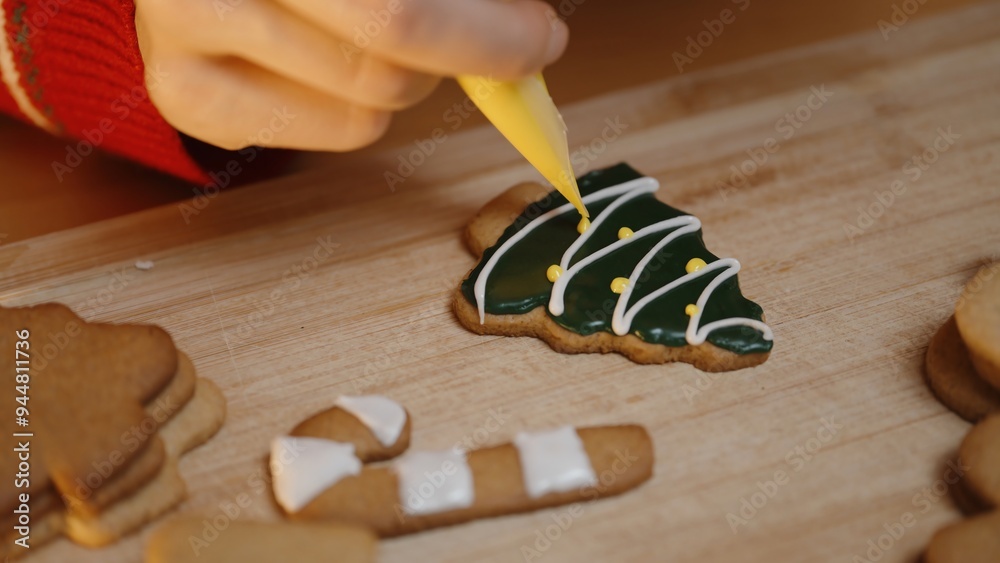 Close-Up of a Hand Adding Yellow Icing Dots to a Green Christmas Tree Cookie With White Icing Stripes on a Wooden Board. Close-Up of a Hand Adding Yellow Icing Dots to a Green Christmas Tree Cookie With White Icing Stripes on a Wooden Board.