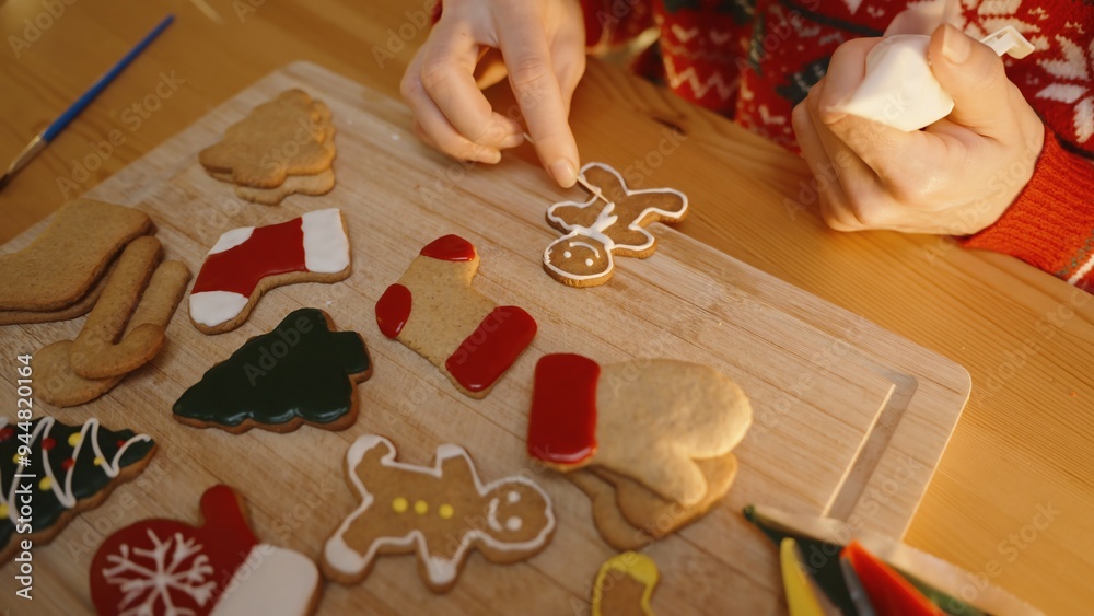 Woman Adds Final Touches to a Gingerbread Man Cookie, Surrounded by Colorfully Decorated Christmas Cookies on a Wooden Board. Woman Adds Final Touches to a Gingerbread Man Cookie, Surrounded by Colorfully Decorated Christmas Cookies on a Wooden Board.