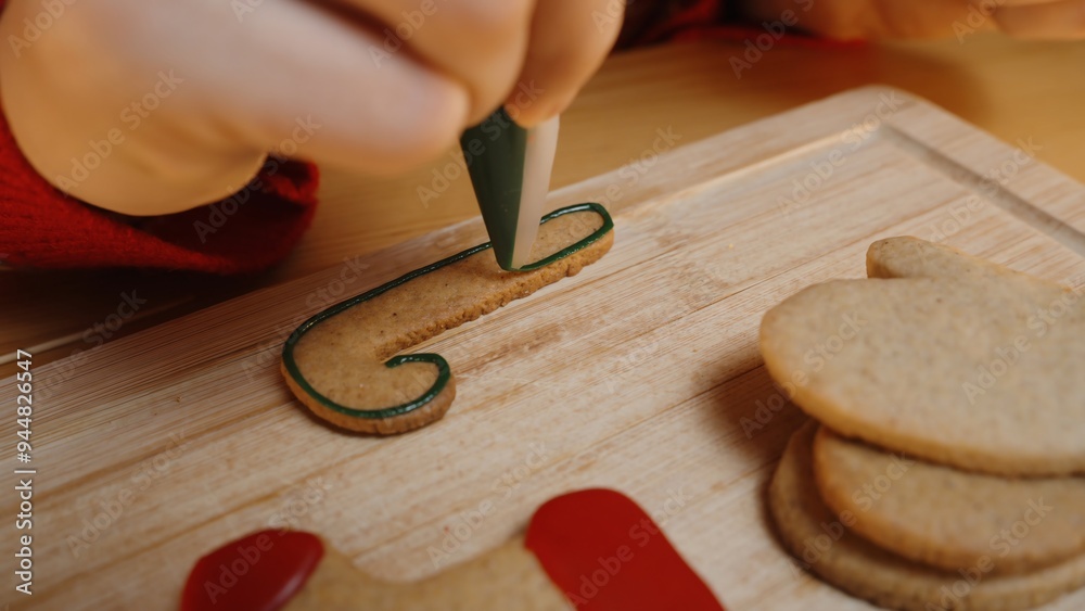 Close-Up of Woman Outlining a Candy Cane Cookie With Green Icing, Preparing to Decorate on a Wooden Board. Close-Up of Woman Outlining a Candy Cane Cookie With Green Icing, Preparing to Decorate on a Wooden Board.