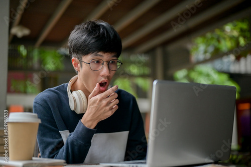 An amazed Asian man looking at his laptop screen, checking aa exam results, or reading shocking news photo