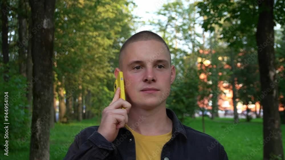 Caucasian young man in casual clothes with a yellow mobile phone. A guy walks alone in the park at sunset. Caucasian young man in casual clothes with a yellow mobile phone. A guy walks alone in the park at sunset.