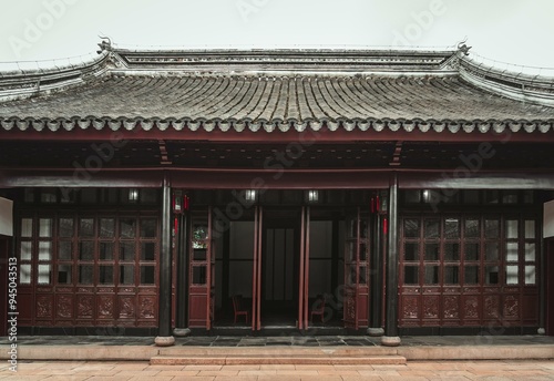 A traditional Chinese building with a curved roof and intricately carved wooden doors. The entrance leads to a courtyard with chairs and a stone floor.