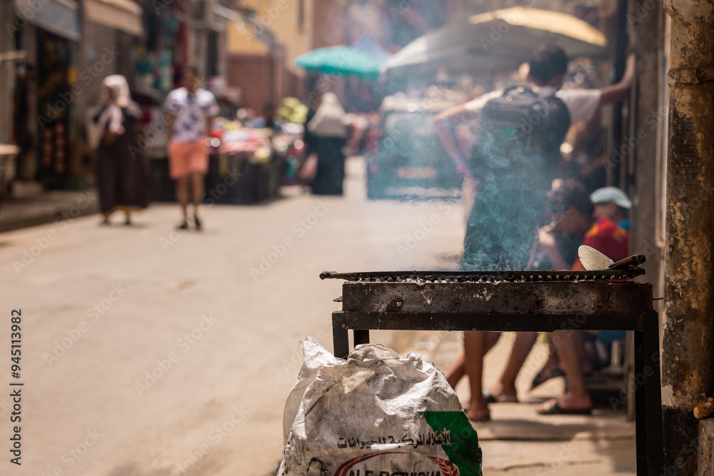 Coal grill being fired up on the market streets of Oran, Algeria. side view of a commercial grill at a market in arab country. Bag of coal next to it, people behind