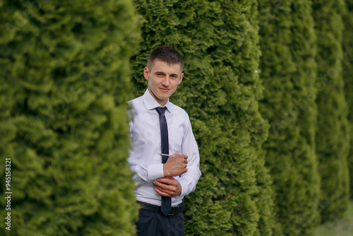 A man in a white shirt and black tie stands in front of a hedge. He is smiling and he is enjoying the moment