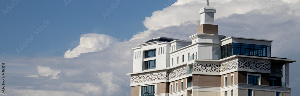 Fragment of the building's architecture against the background of blue sky with white clouds. Combination of complex architectural form and smooth lines of white clouds in the sky. Fragment of the building's architecture against the background of blue sky with white clouds. Combination of complex architectural form and smooth lines of white clouds in the sky.