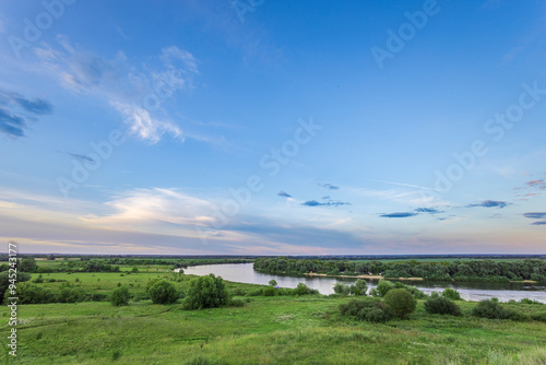 View from hilltop: vast river, lush green meadow, trees on banks. Blue sky, puffy white clouds, warm sunset glow